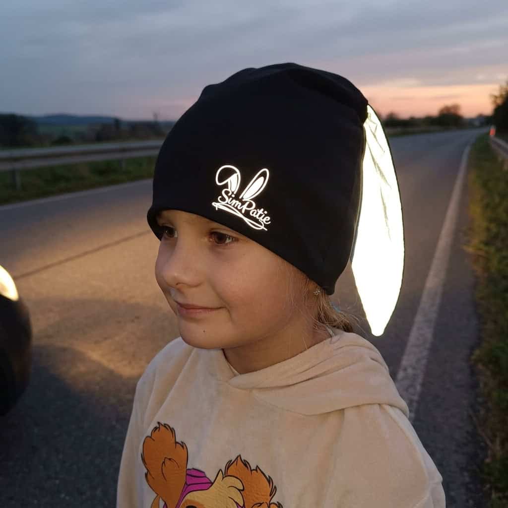 Dítě s černou reflexní čepičkou SimPatic u silnice za šera – reflexní ouška a logo svítí
